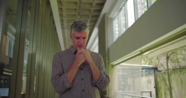 Orta yaşlı bir adam ofis koridorunda, iki elini de yan yana tutarak aşağıya bakıyor. Şirket çevresindeki duygusal endişe ve endişeyi gösteriyor.