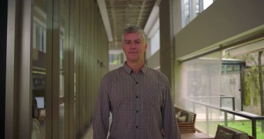 Orta yaşlı bir adam koridorda duruyor. Yumuşak bir gülümsemeyle ofisin iş yeri koridorunda geniş pencereli rahatlamış bir profesyonel ifadeyle.