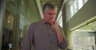 Orta yaşlı bir adam ofis koridorunda elleri ağızlarına yakın duruyor endişeli ifadeye bakıyor profesyonel ortamdaki endişe ve endişeleri iletiyor.