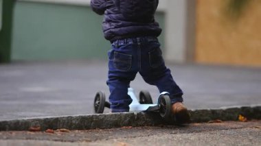 Yürümeye başlayan bir çocuk, sakin bir şehir ortamında erken denge ve hareketi keşfederken, sakin bir kaldırımda scooter sürer ve ileri doğru yumuşak hareketler yapar.
