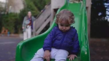 Küçük çocuk yeşil bir oyun alanı kaydırağının ucunda oturur, yumuşak, içe dönük bir ifadeyle aşağıya bakarken hafifçe öne eğilir. Oyunda bir duraksama yakaladığı an çocukluk döneminin hassas duygusal ritmini ve sessiz merakını gözler önüne seriyor.