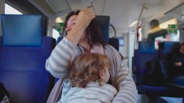 Anne kucağında bebekle trende oturuyor ve gergin bir anında alnını ovalıyor. Yolculuk yorgunluğu ve aile yolculuğundaki bakım sorumluluğunun karışımını gösteriyor.