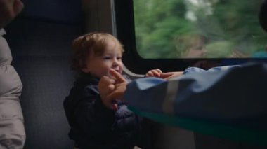Küçük bir çocuk trene binerken gülümser ve güler, küçük bir masada bir kardeşe doğru uzanır. Canlı bir aile etkileşimi, hareket ve basit bir seyahat keyfi yakaladığı an.