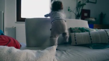 Yeni yürümeye başlayan bir çocuk kanepede duruyor ve aşağıya odaklanmış bir merakla bakıyor, çevreyi erken denge ve dikkatle keşfediyor.