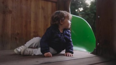 Küçük bir çocuk kaydırağın yanındaki ahşap platformda oturuyor, elleri ve dizleri üzerine çökmeden önce dışarıya bakıyor ve sessiz, neşeli bir gülümsemeyle emekliyor.. 