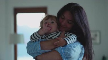 Anne aydınlık evin içinde bebeğe sarılırken çocuk yumuşak bir hareketle dışarıya uzanır. Sıcak sevgiyi, doğal rahatlığı ve aile ortamında duygusal bağı yakalar.