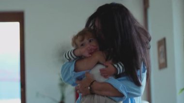 Çocuğunu omzuna almış bir anne, sakin ve güven dolu bir ifadeyle yukarıya bakıyor. Huzurlu ev ortamında duygusal güven ve sıcak aile bağlarını gösteriyor.