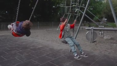 Küçük bir çocuk, büyük bir kardeş yanında sallanırken öne doğru sallanıyor, her ikisi de hareketli kavisler çiziyor. Sahne paylaşılan oyun, hareket ve oyun parkının hareketli ritmini yakalar..