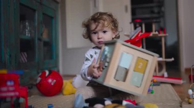 Oyuncaklarla çevrili bir şekilde oturan küçük çocuk, sessiz oyun zamanı boyunca doğal konsantrasyon ve merakı gösteren ahşap bir aktivite kutusunun parçalarıyla deney yapıyor.