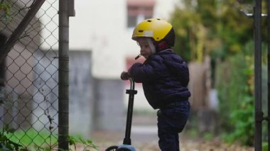 Yürümeye başlayan bir çocuk, yeşillik ve zincirli tel örgüyle çevrili dar bir açık yolda ilerlemeye devam etmeden önce bir scooter 'da durup merakla bakar.