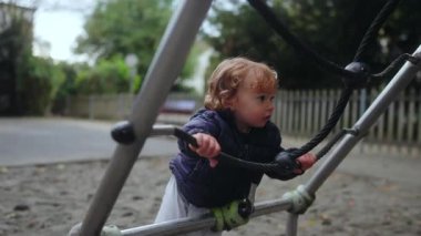 Bir çocuk halat yapısına tutunarak tırmanmaya çalışır, kum ve sessiz bir park zeminiyle çevrili oyun alanını keşfederken erken çaba ve merakını gösterir.