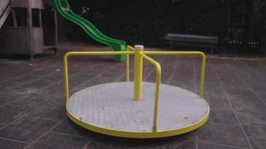 Sarı bir döner kavşak, yeşil bir kaydırak, ahşap bir oyun yapısı ve sakin asfalt bir alan ile çevrili, boş bir oyun alanında döner nazik bir hareket ve açık bir çevre gösterir