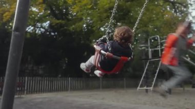 Küçük çocuk güvenli koltukta sallanırken büyük kardeş kendinden emin bir şekilde onun yanında sallanarak hareketli bir hareket ve erken bağımsızlık anı yaratıyor..