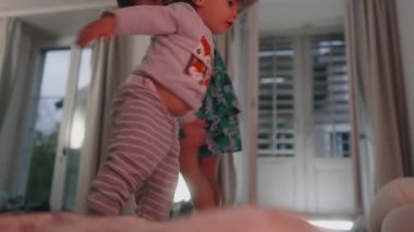 Çocuklar bir yatakta enerjik bir şekilde tırmanıyor ve hareket ediyor, merak, hareket ve rahat bir iç mekan ortamında spontane çocukluk etkileşimiyle dolu neşeli bir an yaratıyorlar.