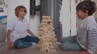 İki çocuk tamamlanmış ahşap kuleye doğru heyecanlı bir gülümsemeyle oturur, yaratıcılık, takım çalışması ve birlikte bir şeyler inşa etmenin sevinci ile dolu sıcak, neşeli bir an oluştururlar.