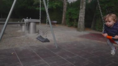 Küçük bir çocuk salıncakta ileri geri sallanıyor, yumuşak, sessiz bir ifadeyle yana bakıyor zincirleri tutarken..