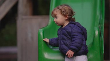 Bir çocuk yeşil oyun parkında sessizce oturur, başını çevirir ve başka bir çocuk onun önünden hızla geçer. Erken sosyal farkındalığı, akranlara olan merakı ve küçük çocukların gözlemsel doğasını yansıtıyor.