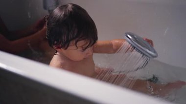 Bir çocuk küvette oturmuş duş başlığını inceliyor, küvete su sıçrayan akıntıyı seyrediyor. Sakin an, merakı, duyusal oyunu ve erken duş alma bağımsızlığını vurguluyor..