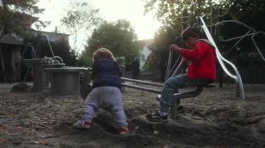 Küçük bir çocuk kum kazıcı kolunun altındaki metal bağlantı çubuğuna uzanırken ağabeyi de yakınlardaki kumu temizlemeye devam ediyor. Etrafı parkın tadını çıkaran diğer çocuklarla çevrili küçük çocuk kendini yumuşak zemine sabitlemeye çalışıyor.