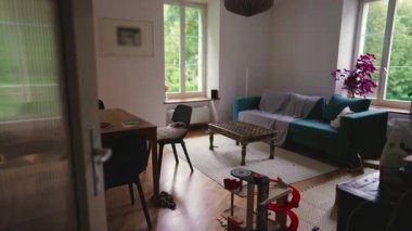 Geniş pencereli parlak bir oturma odası, bir kanepe, çocuklar için oyuncaklar, ve bir masa hoş bir ev ortamı yaratır. Mekan, günlük aile yaşantısını ve rahat bir çevreyi yansıtıyor..