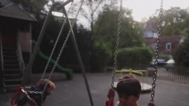 Bir çocuk havada yükselir salıncakta, bebek yavaşça aşağı doğru hareket ederken hareketli dinamik bir kontrastı ve açık havada oyun oynamanın ortak sevincini yakalar.