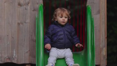 Bir çocuk yeşil oyun alanı kaydırağını indirir, her iki tarafı da denge için tutar ve dikkatlice aşağıya doğru hareket eder. Erken kendine güveni, hafif heyecanı ve çocukluk oyununun basit neşesini yakaladığı an harekete geçer.