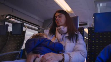 Anne, bebeğini sessiz bir trende emziriyor. Kırsal alanda dolaşırken şefkat ve yorgunluk karışımı bir şey gösteriyor. Uzun bir yolculukta samimi ve sakin bir aile anı yaratıyor.