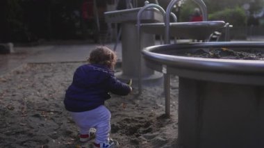 Yeni yürümeye başlayan bir çocuk, açık hava oyununun bir parçası olarak havaya bir kum patlaması göndererek sarı bir plastik kepçe sallıyor. Saf çocukluk sevincini, duyusal bağlılığı ve açık oyun alanlarında ortaya çıkan maceraperest merakı yakalar..