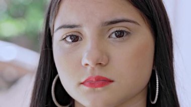 Ciddi bir ifadeyle kameraya bakan, küpe takan, ruj süren, arka planda bulanık duran genç bir kız.