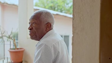 Duvara yakın oturan yaşlı bir adamın yan profili, yumuşak bir ifade ve rahat bir dış koridor ayarı ile