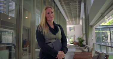 Koridorda duran bir kadın, göğsünü ve tabletini sıkıca tutuyor, iş ortamındaki gergin anlarda rahatsızlık veya duygusal hassasiyet gösteriyor.