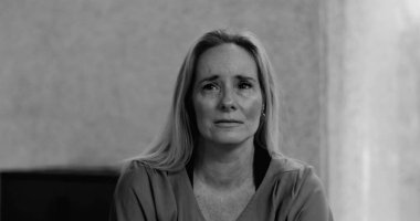 Kadın doğrudan kameraya bakıyor gözyaşları içinde derin üzüntü ve kırılganlık ifade ederken sessiz, samimi bir şekilde içeride oturuyor, siyah ve beyaz.