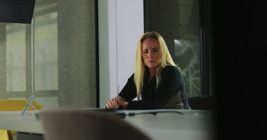 Kadın, modern ofis ortamında masa aksesuarları, dizüstü bilgisayarlar ve sandalyelerle çevrili, düşünceli bir şekilde konferans masasında oturuyor.