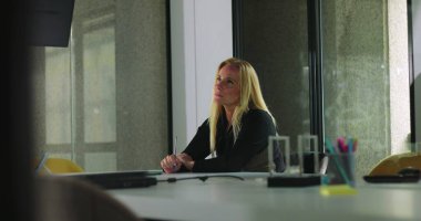 Kadın, modern ofis ortamında masa aksesuarları, dizüstü bilgisayarlar ve sandalyelerle çevrili, düşünceli bir şekilde konferans masasında oturuyor.