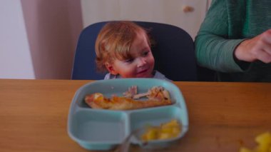 Yeni yürümeye başlayan çocuk, elinde bölünmüş bir tabak yiyecekle yemek masasında oturmuş, evde normal bir aile yemeği sırasında yanında yemek hazırlayan ya da kesen ebeveyne bakıyor..