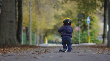 Yürümeye başlayan çocuk, ağaçlarla çevrili yaprak kaplı bir patika boyunca arka planda park işaretleri ve uzak sokak ayrıntılarıyla birlikte giderken kameraya bakan üç tekerli bir scooterla ileri doğru hareket ediyor.