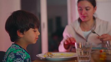 Anne, yemek sırasında masaya tabak, kap kacak ve bardaklarla birlikte otururken, küçük çocuğa bir lokma yemek yedirmek için masaya uzanır.