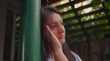 Bir kadın nazikçe yansıtıcı bir ifadeyle elini yüzüne koyar, açık hava ışığı ve hafif bir esinti gibi yavaşça bir direğe yaslanır sıcak bir iç gözlem atmosferi yaratır.