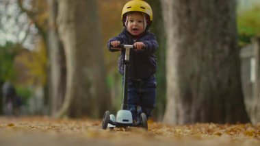 Yürüyen çocuk, gidonu tutarak ve arka planda büyük ağaçlar ve park çevreleri olan yapraklarla kaplı bir yolda scooter 'ı ileri doğru yürüterek ilerler.