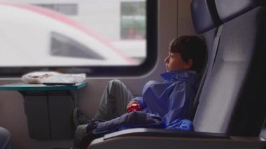 Bir çocuk koltukta dinlenen bacakları ile trende rahatça oturur, sessizce bulanık manzaranın pencerenin önünden geçişini izler. Araziyi sakin bir ilgiyle gözlemler, yolculuk sırasında huzurlu ve içsel bir an yaratır..