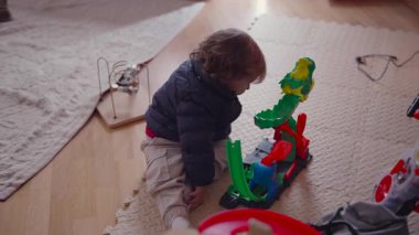 Bebek oturma odasındaki yastıklı bir paspasın üzerine oturmuş renkli plastik bir oyuncak pistini keşfetmeye odaklanmış. İçerde sessizce oynarken parçalara dokunur ve oyuncakla etkileşime girer..