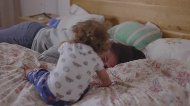 Anne ve bebek rahat bir sabah rutini sırasında yatakta şakalaşarak, huzurlu ev atmosferinde sevgi, güven ve doğal bağlılıkla dolu hakiki bir aile anı yaratıyorlar.