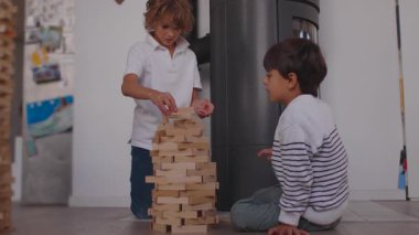 İki çocuk birlikte oturup, yuvarlak ahşap bir kuleyi şekillendiriyor, dikkatli hareketlerle bloklar ekliyor ve kapalı alandaki oyun sırasında sessizce paylaşılan konsantrasyonu sağlıyor.