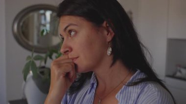 Mutfakta eli ağzının yanında duran bir kadın, düşüncelere dalmış bir şekilde duraklarken odadan gelen ışık onun düşünceli yüz ifadesini vurguluyor, her gün yansıtıcı bir anı yakalıyor..