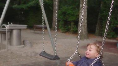 Yürüyen bebek kova salıncakta oturuyor. Daha büyük çocuk oyun alanı elemanlarıyla ileri doğru hareket ediyor. Mesela sürgülü adımlar ve açık hava parkında görünen metal çerçeve gibi.