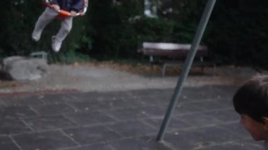 Küçük çocuk salıncakta sallanırken büyük çocuk salıncak zinciri ve çerçevenin etrafında açık park alanı ile hareketlere rehberlik etmek için koltuğun ön tarafını tutuyor.