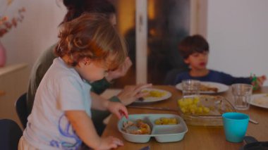 Küçük çocuk yemek masasında elleriyle yiyecek ararken anne ve kardeşi yakınlarda yemek yiyor ve paylaşılan aile yemeğine özerklik merakını ve dahil edilmeyi ifade ediyor.