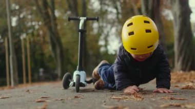 Yürüyen çocuk, üç tekerli scooter 'dan düştükten sonra, yapraklarla kaplı park yolunda oturup şikayet ediyor, scooter yakınlarda duruyor ve kaldırım boyunca büyük ağaçlar görünüyor.