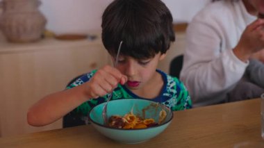 Genç çocuk yemek masasında çatalla makarna yerken bulaşıklar, çatallar ve bardaklar tipik bir ev yemeği ortamında masada duruyor.