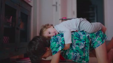 İki çocuk, oturma odası mobilyaları ve oyuncaklarıyla evin arka planında görünür bir şekilde etkileşime girerken, küçük kız kardeş yerde neşeli bir anda esniyor.